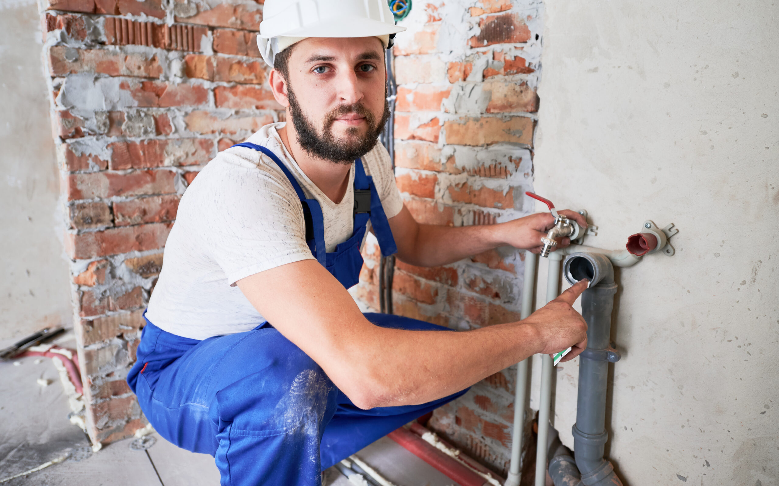 Handsome male worker in safety helmet looking at camera while applying silicone grease on pipe. Man plumber in work overalls lubricating pipe while installing water system in apartment.